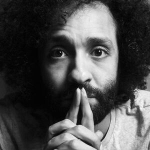 Black and white portrait of YKY Sadek, gazing thoughtfully with hands clasped near his face. He has curly hair and a contemplative expression.